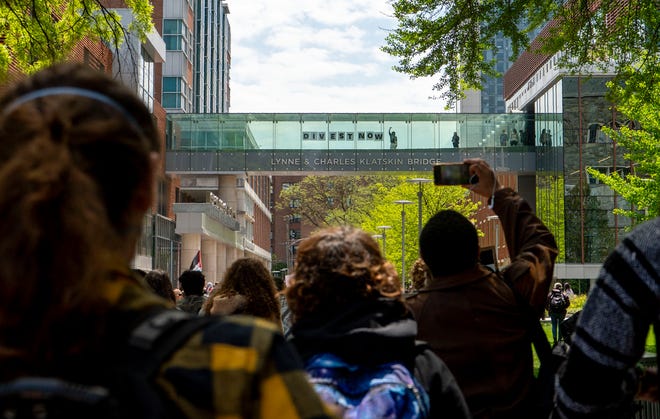 A pro-Palestinian protester stands on the Lynne and Charles Klatskin Bridge at Temple University with a sign reading "Divest Now" on April 25, 2024, as the Students for the Justice in Palestine and Philly Palestine Coalition members and supporters march toward them on campus in Philadelphia to demand the university divest from companies with ties to the Israeli government.